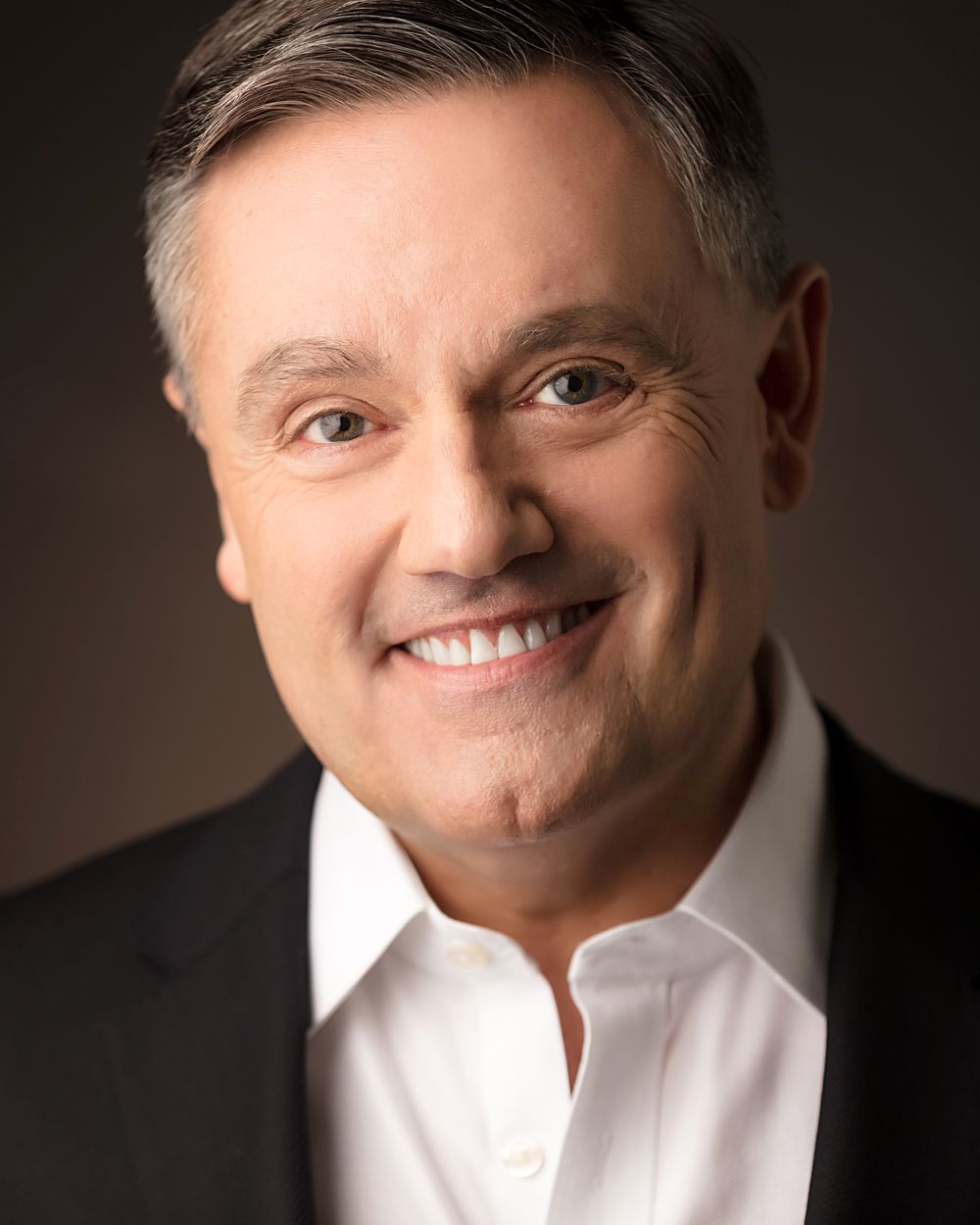 Commercial actor headshot with a warm smile, dark suit and white shirt on a brown backdrop, HeadshotDC