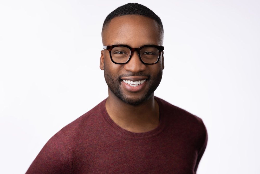 Portrait of a smiling young man wearing glasses and a maroon sweater against a white background.