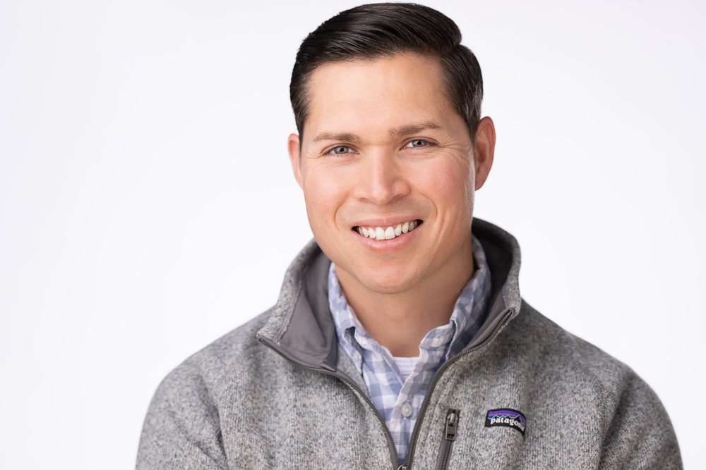 A smiling man in a gray Patagonia jacket and blue checkered shirt against a white background.