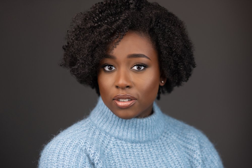 A portrait of a young African American woman with curly hair wearing a light blue sweater against a dark background.