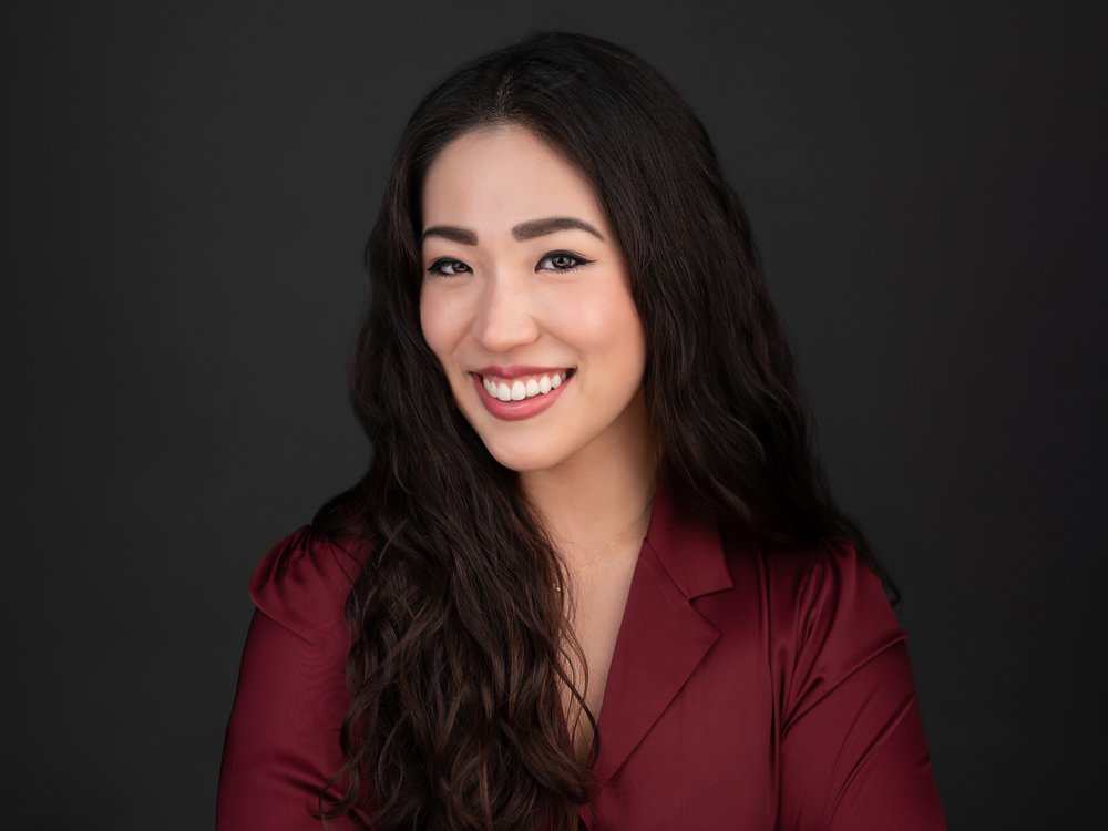 A young woman with long dark wavy hair, smiling, wearing a burgundy blouse, against a dark background.