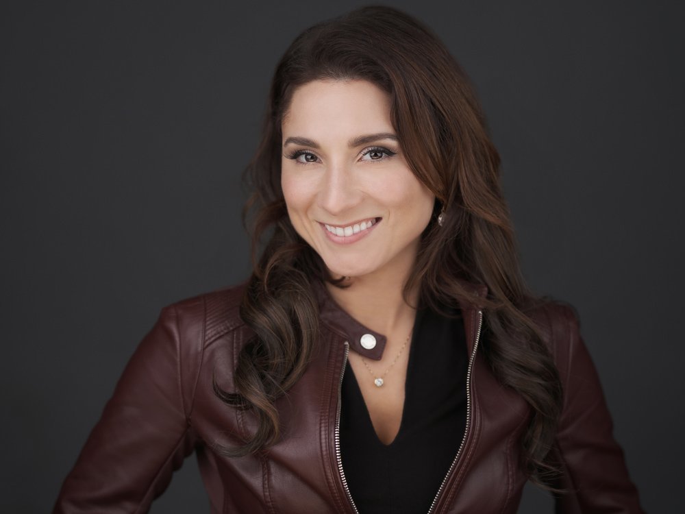 A woman with brown hair and a smile, wearing a dark top and a brown leather jacket against a dark background.