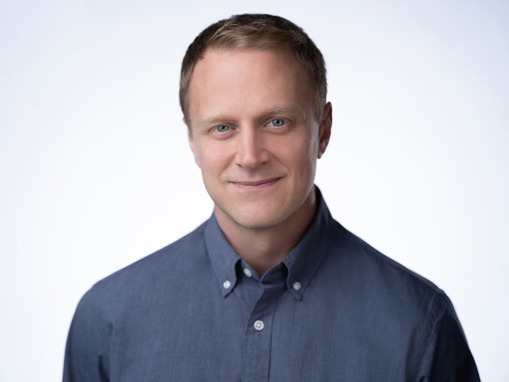 Headshot of a man with short light brown hair, blue eyes, wearing a dark blue button-up shirt, smiling against a plain white background.