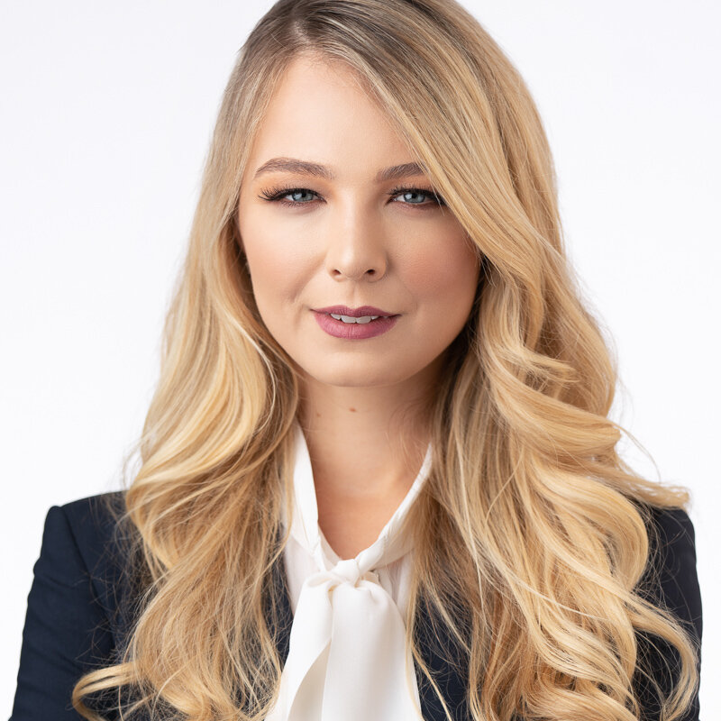 A woman with long blonde hair, wearing a black blazer and white blouse, looking directly at the camera with a slight smile.
