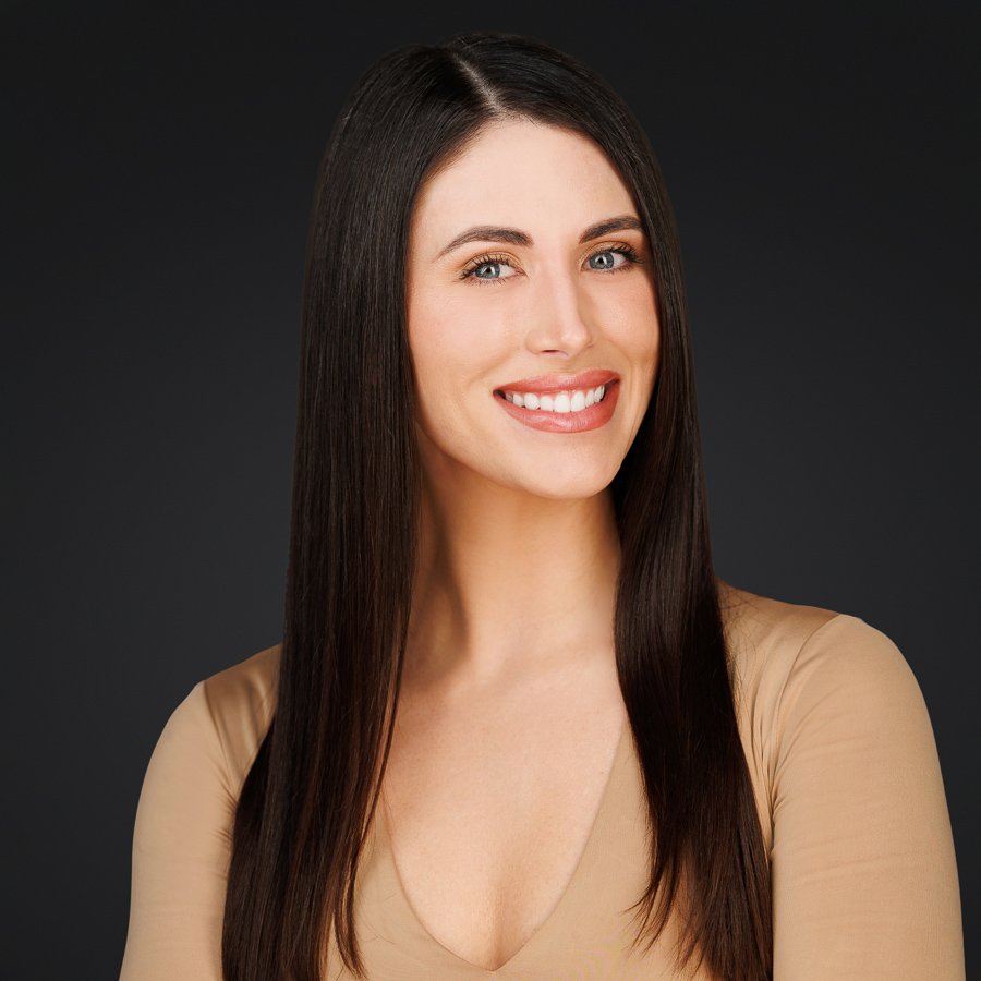 A woman with long, straight brown hair smiling and wearing a beige top against a dark background.
