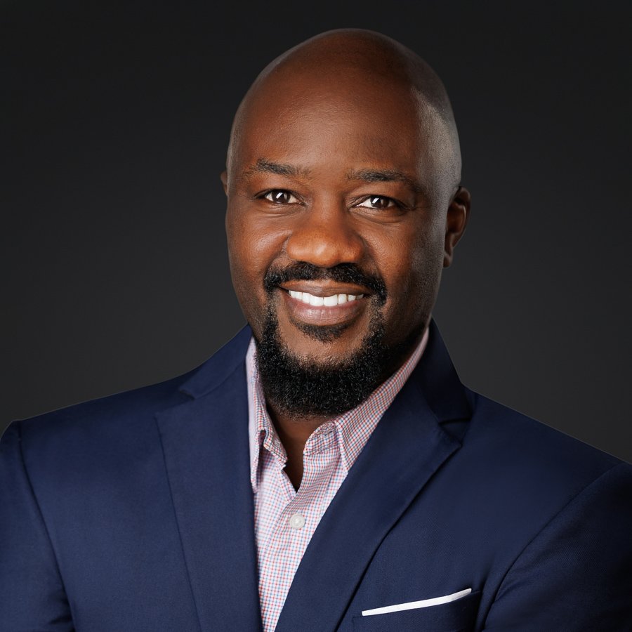Headshot of a smiling African American man with a bald head and a beard, wearing a navy blazer and a checkered shirt, against a dark gray background.