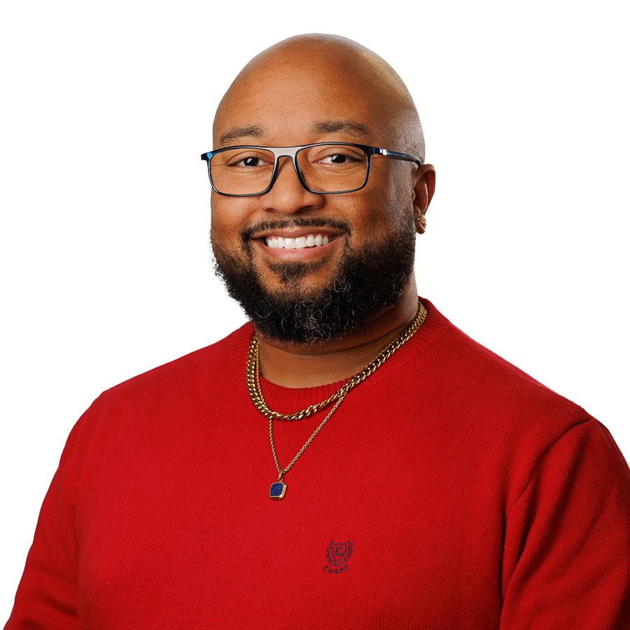 A smiling man with glasses, a beard, and earrings, wearing a red sweater and gold necklaces posed against a white background.
