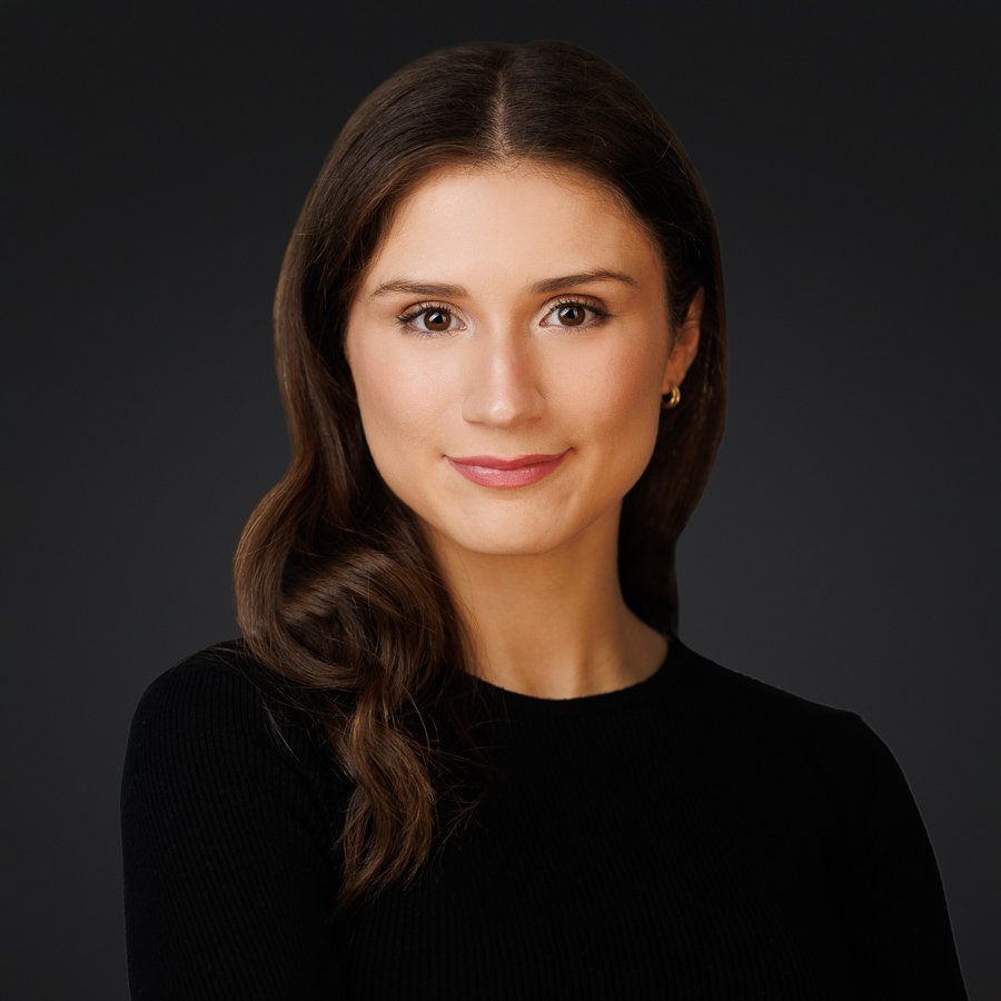 Headshot of a woman with long brown hair, fair skin, wearing a black top, smiling, against a dark background.
