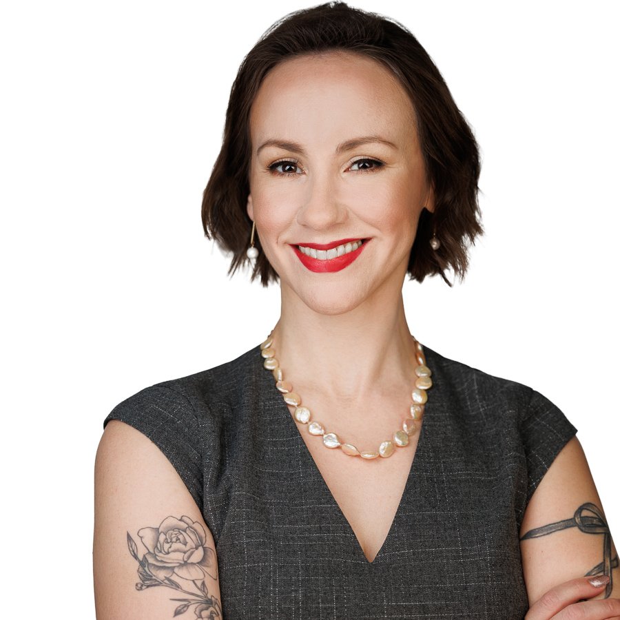 Portrait of a woman with short brown hair, wearing a dark gray dress, pearl necklace, and pearl earrings, smiling and winking, with tattoos on both arms, against a white background.