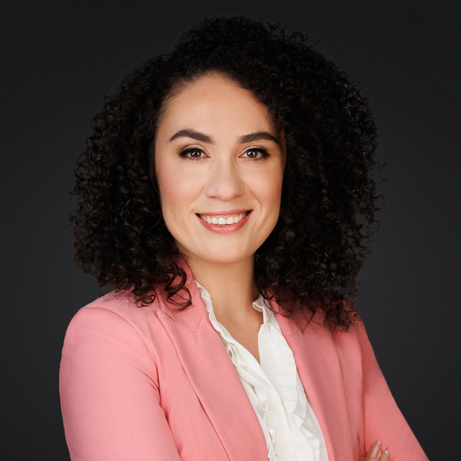 A woman with curly dark hair, wearing a pink blazer and white blouse, smiling against a dark background.