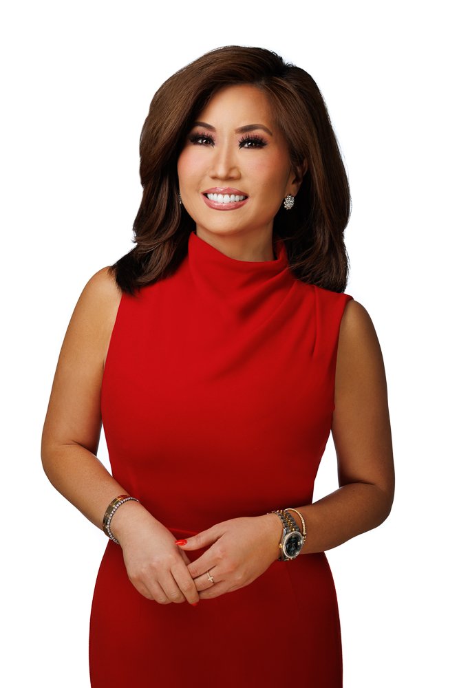 A woman with shoulder-length brown hair, wearing a sleeveless red dress, and accessorized with jewelry including earrings, bracelets, and a watch, standing against a white background.