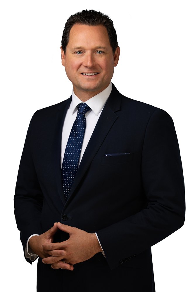 A man in a dark suit with a white shirt and a blue dotted tie standing against a white background, smiling with hands clasped in front of him.