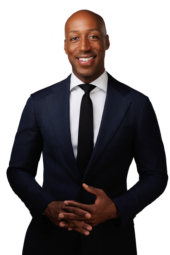 A smiling African American man in a dark suit with a white shirt and black tie, standing with hands clasped in front of him, against a white background.