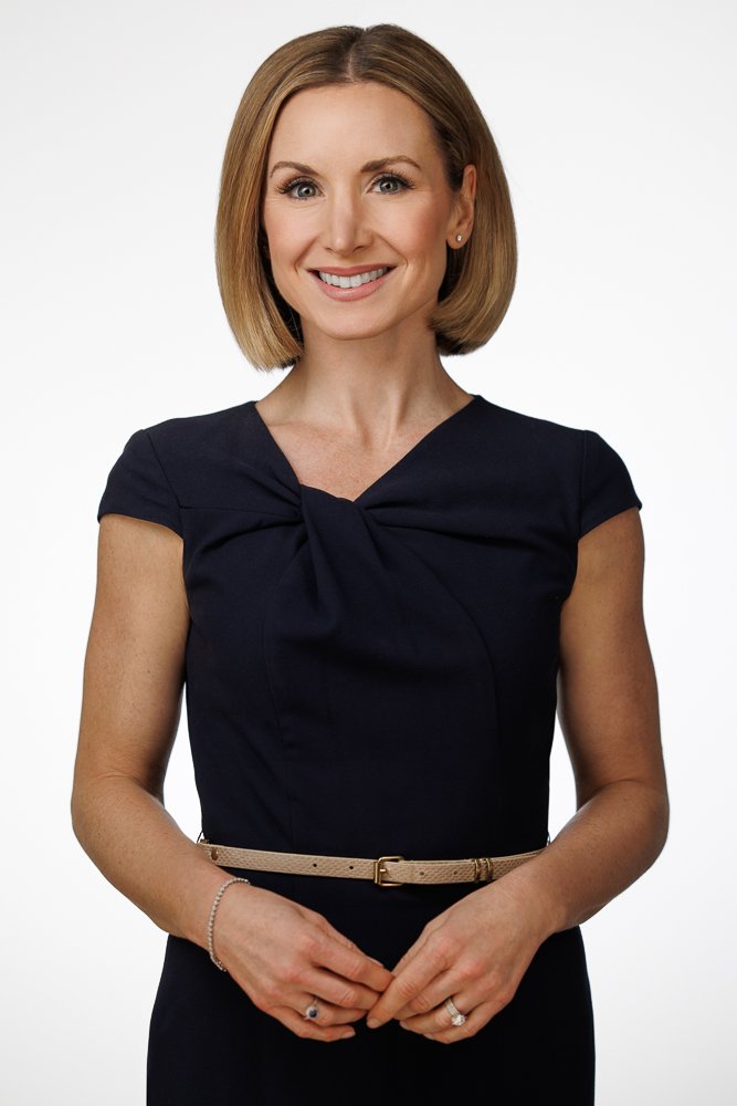 A woman with shoulder-length blonde hair, wearing a dark navy dress with a bow detail on the front, standing against a white background, smiling with hands clasped in front.
